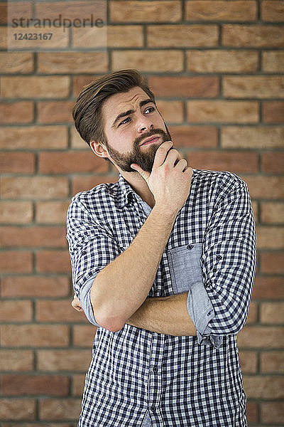 Portrait of bearded young man in front of brick wall thinking