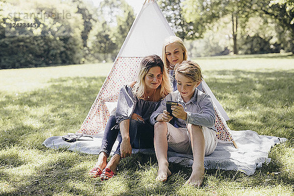 Two young women and a boy looking at cell phone next to teepee in a park