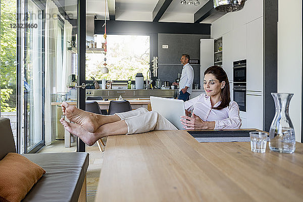 Relaxed woman at home using a laptop at table with man in background