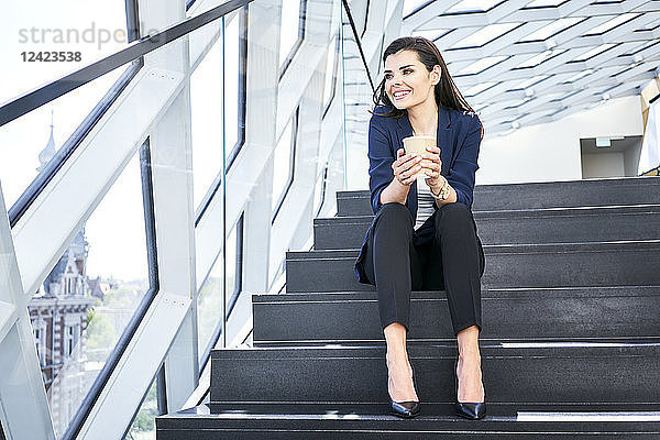 Smiling businesswoman sitting on stairs having a coffee break in modern office