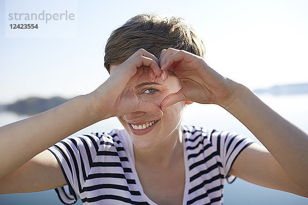 Portrait of smiling woman in front of lake shaping heart with her fingers