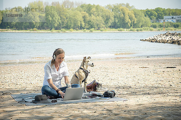 Woman sitting on blanket at a river with dog wearing headphones and using laptop