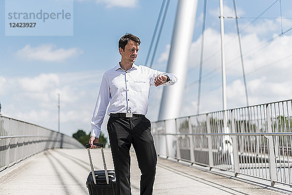 Businessman with rolling suitcase on a bridge checking the time