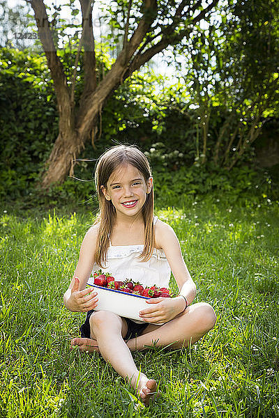 Portrait of smiling little girl sitting on meadow with bowl of strawberries