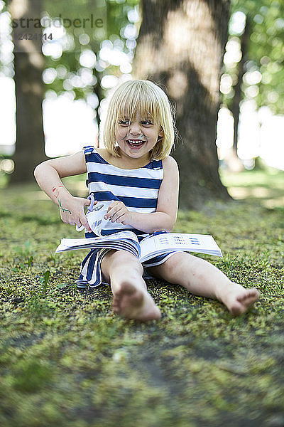 Portrait of laughing little girl with painted face sitting on ground in nature