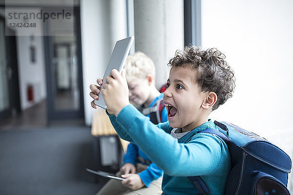Happy schoolboy holding up tablet on corridor in school