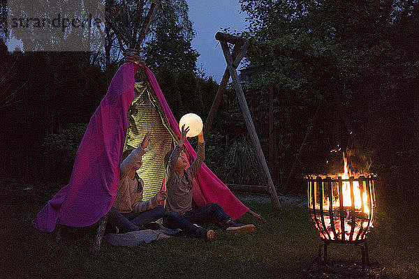 Two girls standing in front of tipi  holding lamp as moon