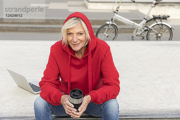 Senior woman wearing red hoodie sitting outdoors with laptop and takeaway coffee