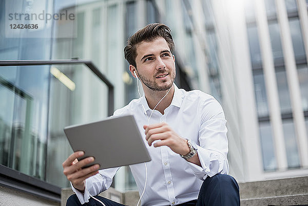 Smiling businessman sitting on stairs with earbuds and tablet