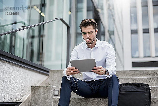 Businessman sitting on stairs with earbuds and tablet