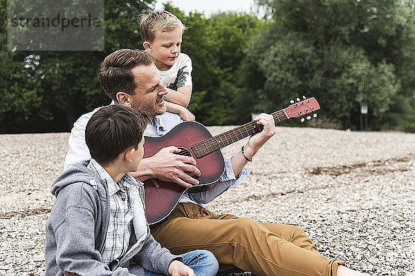 Father with two sons sitting on pebble beach playing guitar