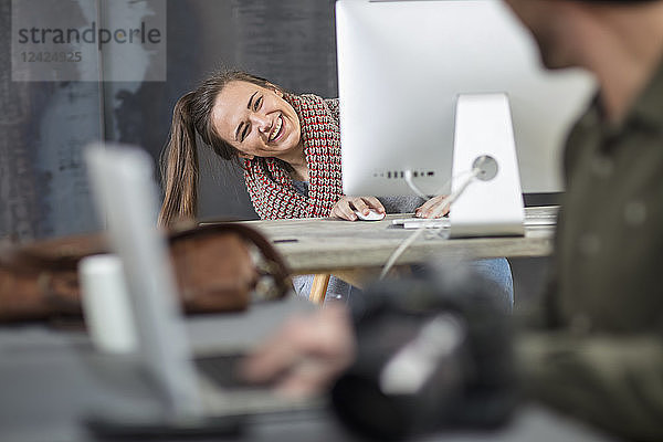 Happy young woman peeking around computer screen in office