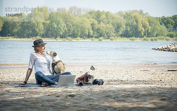 Woman sitting on blanket at a river with dog and laptop