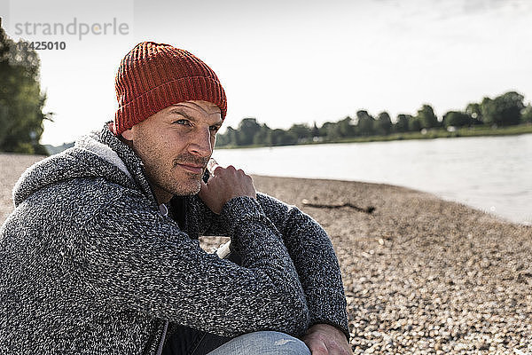 Mature man wearing red beanie sitting at Rhine riverbank
