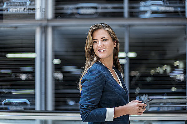 Portrait of smiling young businesswoman with cell phone in front of blurred parking garage