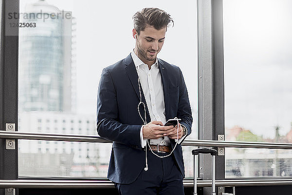 Young businessman at the window with cell phone  earbuds and rolling suitcase