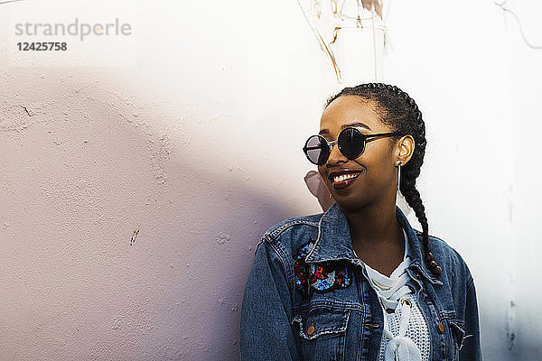 Lächelnde junge Frau mit Sonnenbrille und Jeansjacke vor einer weißen Wand