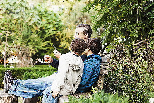 Vater und zwei Söhne (10-11) sitzen auf einer Bank im Park und schauen auf ihr Smartphone