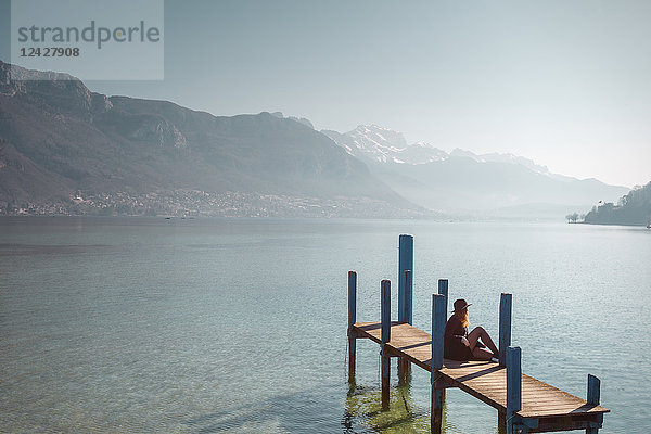 Frau sitzt auf einem Steg am Seeufer unter klarem Himmel mit Bergen im Hintergrund  Annecy  Haute-Savoie
