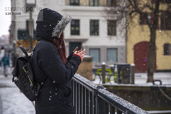 Seitenansicht einer Frau mit Kapuzenjacke und Rucksack  die ein Foto mit ihrem Smartphone auf einer Brücke in Kopenhagen  Dänemark  macht