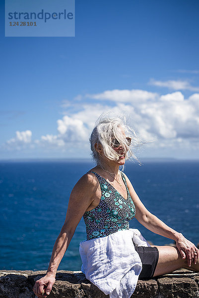 Reife Frau mit grauem Haar sitzt gegen das Meer unter blauem Himmel und Wolken