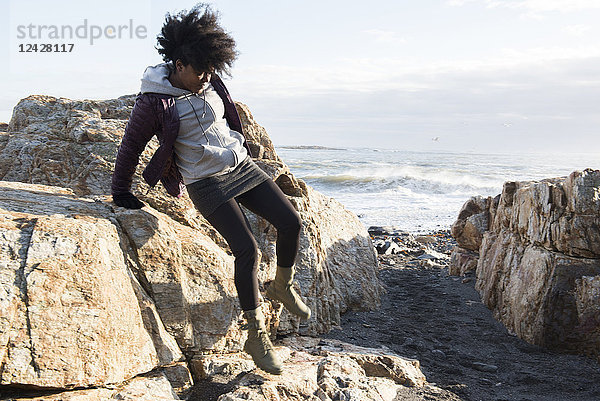 Eine afroamerikanische Frau in einer lila Jacke springt von einem Felsen am Meer herunter
