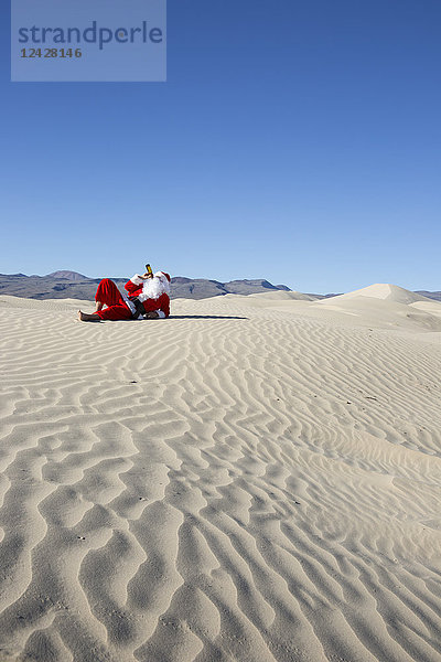 Weihnachtsmann trinkt Bier  während er auf Sand in der Wüste unter klarem Himmel liegt