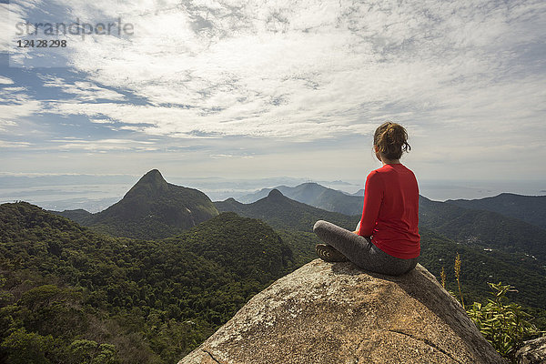 Rückansicht einer einzelnen Frau  die auf einem Felsen in einer Landschaft mit Bergen sitzt  Tijuca Forest National Park  Rio de Janeiro  Brasilien