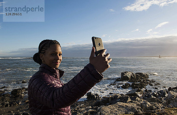 Taillenaufnahme einer alleinstehenden afroamerikanischen Frau  die ein Selfie am Meer macht