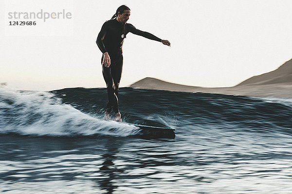Surfer im Neoprenanzug auf einer Welle im Meer bei Sonnenuntergang in voller Länge