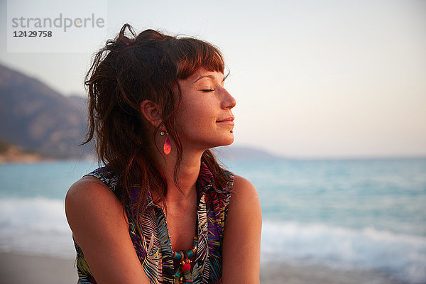 Kopf und Schultern Porträt der schönen jungen Frau mit geschlossenen Augen am Strand bei Sonnenuntergang