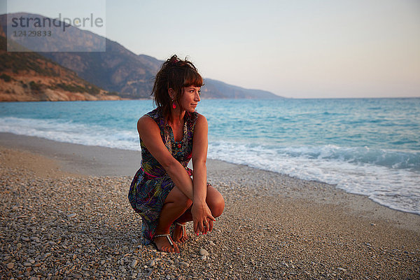 Ganzkörperporträt einer schönen jungen Frau  die bei Sonnenuntergang am Strand kauert