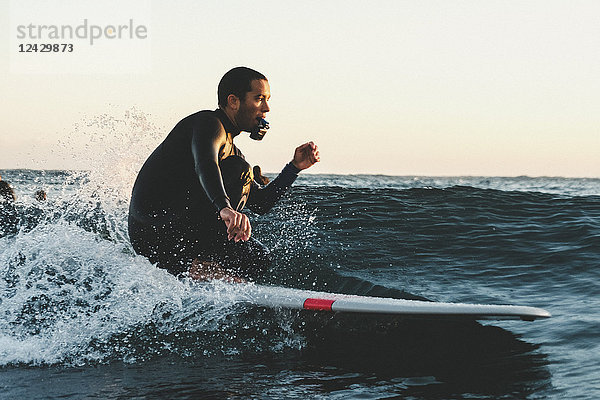 Seitenansicht eines Surfers im Neoprenanzug  der auf einem Surfbrett mit einer tragbaren Kamera im Meer hockt