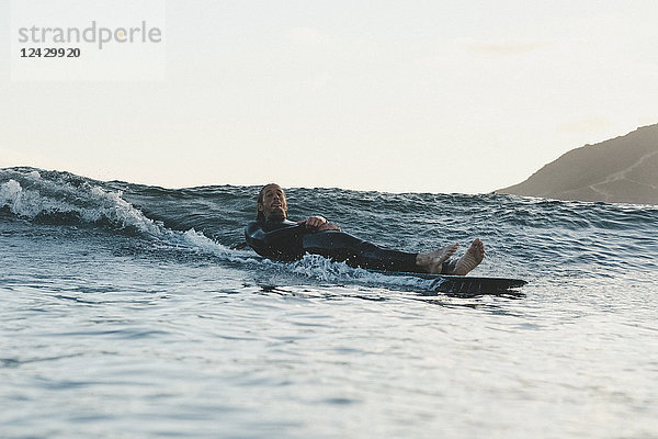 Totale Aufnahme eines Mannes im Neoprenanzug beim Surfen  der auf einem Longboard liegt und in die Kamera schaut