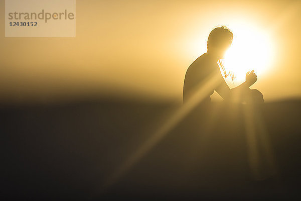 Eine Frau  die auf einem Surfbrett im Meer sitzt  erscheint als abstrakte Figur  die in das goldene Licht des Sonnenuntergangs gehüllt ist