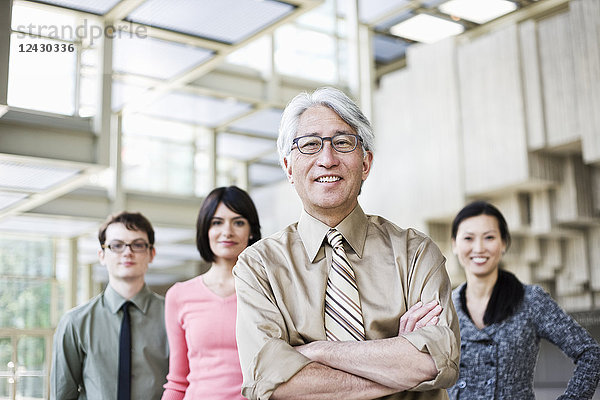 Ein Porträt eines gemischtrassigen Teams von Geschäftsleuten  das im Lobbybereich eines Kongresszentrums mit einem asiatischen Geschäftsmann an der Spitze steht.