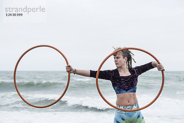 Junge Frau mit braunen Haaren und Dreadlocks  die am Meer steht und zwei Hula-Hoop-Reifen balanciert.