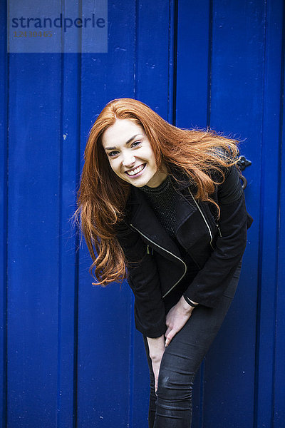 Porträt einer lächelnden jungen Frau mit langen roten Haaren vor einer hellblauen Tür.