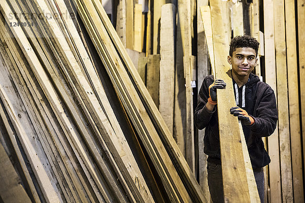 Junger Mann mit Arbeitshandschuhen steht in einem Lagerhaus neben einem Stapel Holzbretter  trägt lange Holzstücke und schaut in die Kamera.
