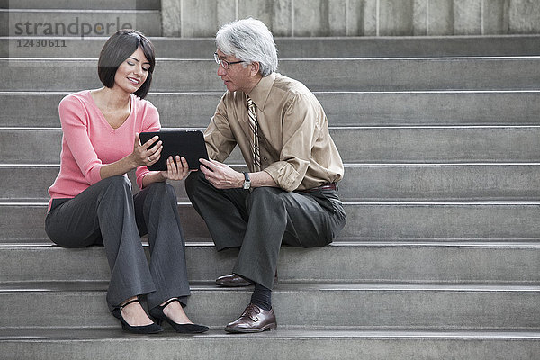 Eine hispanische Geschäftsfrau und ein asiatischer Geschäftsmann arbeiten an einem Notebook  während sie auf den Stufen eines Kongresszentrums sitzen.