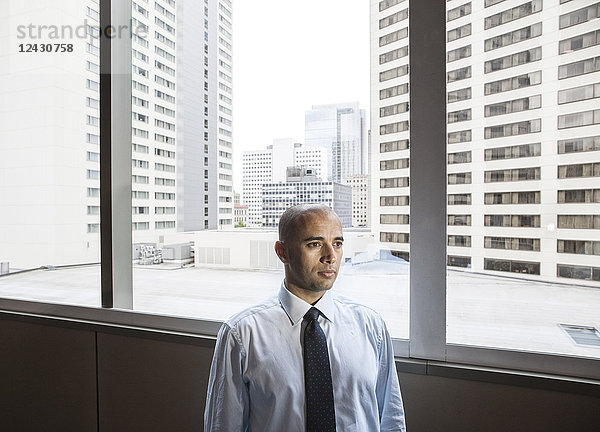 Ein Geschäftsmann aus dem Nahen Osten steht neben einem Fenster  das auf eine Stadtlandschaft mit hohen Bürogebäuden blickt.