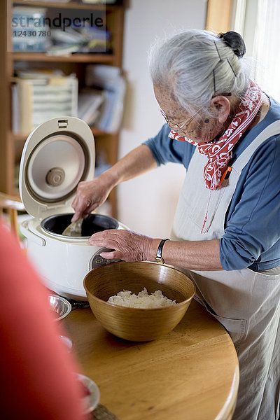 Ältere Frau  die an einem Tisch in einer Küche steht und Sushi-Reis vom Reiskocher in eine Schüssel löffelt.