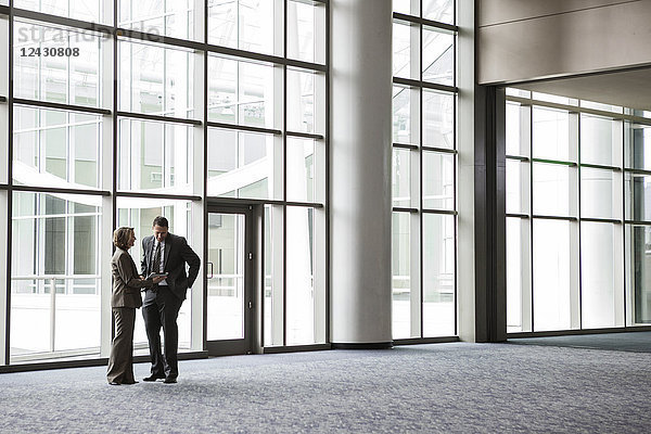 Eine kaukasische Geschäftsfrau und ein Mann bei einem informellen Treffen vor einem großen Fenster in der Lobby eines Kongresszentrums.