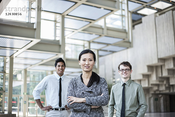Ein Porträt eines gemischtrassigen Teams von Geschäftsleuten  das im Lobbybereich eines Kongresszentrums mit einer asiatischen Geschäftsfrau an der Spitze steht.
