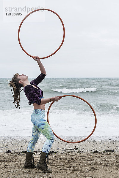 Junge Frau mit braunen Haaren und Dreadlocks  die an einem Sandstrand am Meer steht und zwei Hula-Hoop-Reifen balanciert.