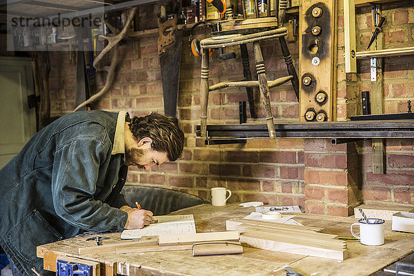Mann arbeitet stehend an einer Werkbank in einer holzverarbeitenden Werkstatt und schreibt in ein Notizbuch.