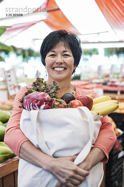 Lächelnde Frau steht in einem Lebensmittel- und Gemüsemarkt und hält eine Einkaufstasche mit frischen Produkten wie Bananen  Tomaten und Kohl in der Hand.