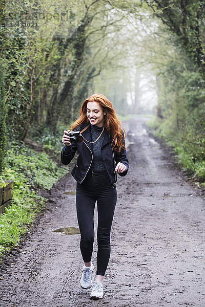 Lächelnde junge Frau mit langen roten Haaren  die einen Waldweg entlang läuft und mit einer Oldtimer-Kamera fotografiert.
