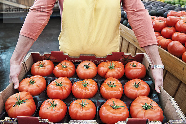 Nahaufnahme aus hohem Winkel einer Person  die auf einem Obst- und Gemüsemarkt ein großes Tablett mit frischen Tomaten hält.
