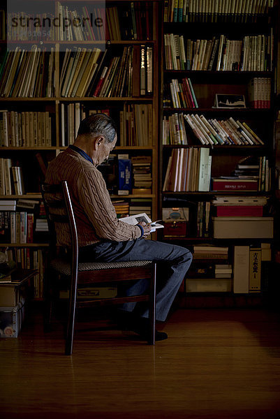 Älterer Mann sitzt auf einem Stuhl vor einem Bücherregal und liest ein Buch.